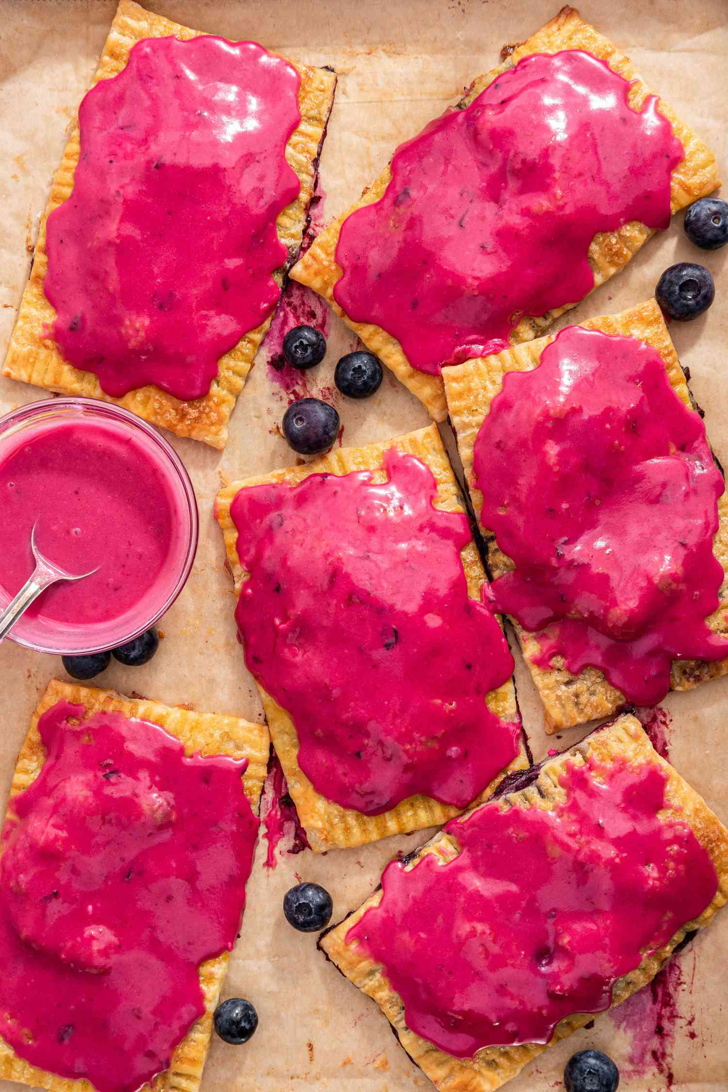 Blueberry Hand Pies on a Parchment Paper Lined Baking Sheet With Bowl of Glaze and Blueberries Sprinkled Around the Hand Pie