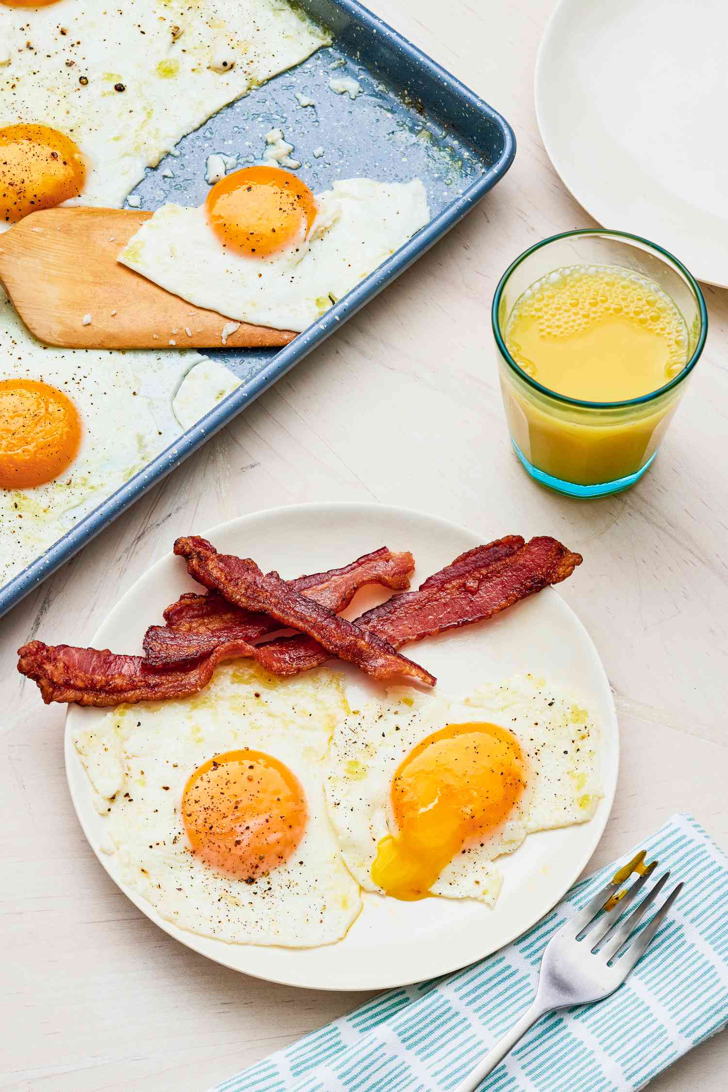 Plate of Sheet Pan Fried Eggs Served With Bacon, and in the Surroundings, a Glass of Orange Juice, a Fork on a Table Napkin, Another Plate, and More Sheet Pan Fried Eggs on a Sheet Pan