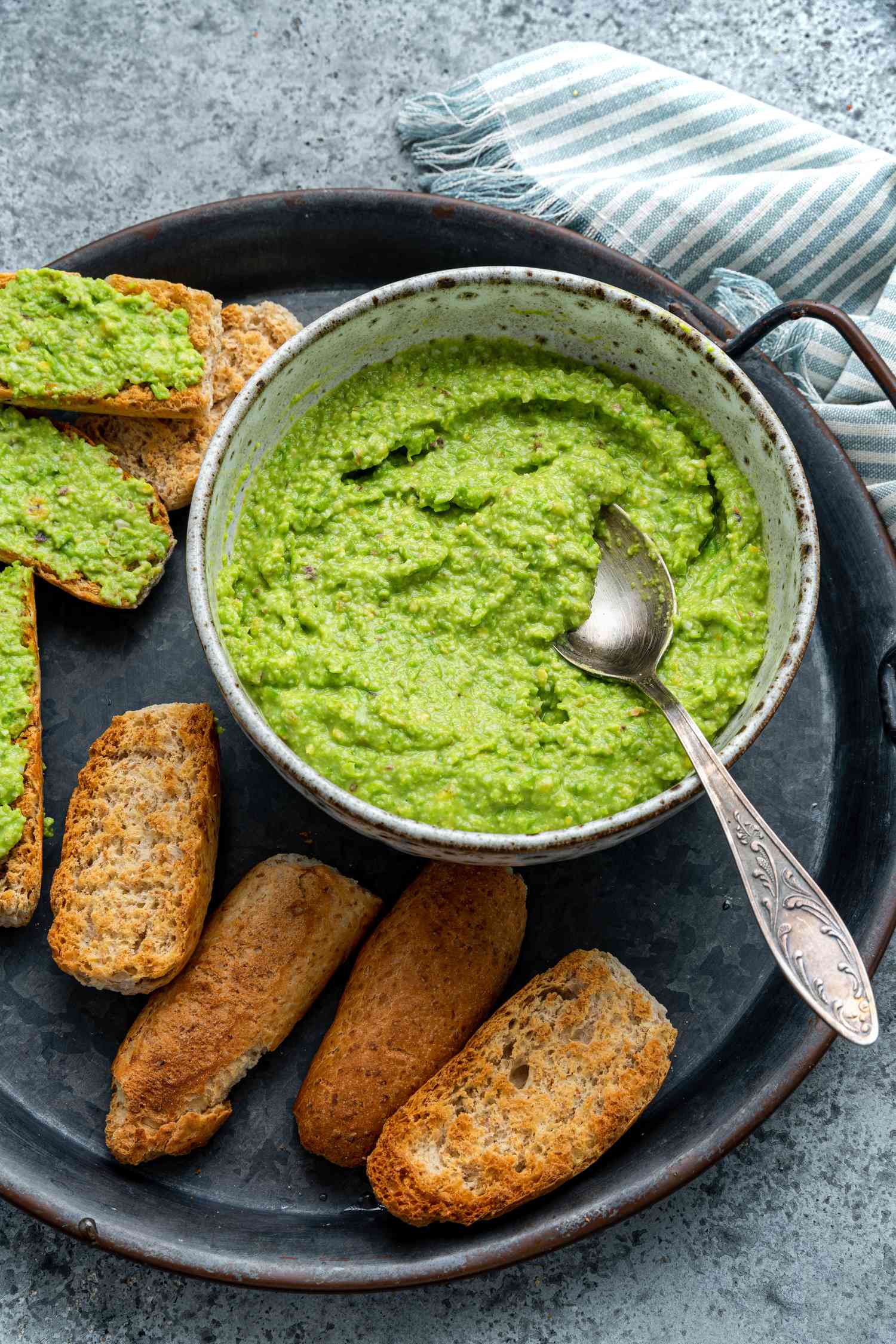 Bowl of Pea Pesto on a Tray With Toasted Pieces of Bread (Some Have Pesto Smeared on It), and Next to the Tray, a Blue and White Kitchen Towel