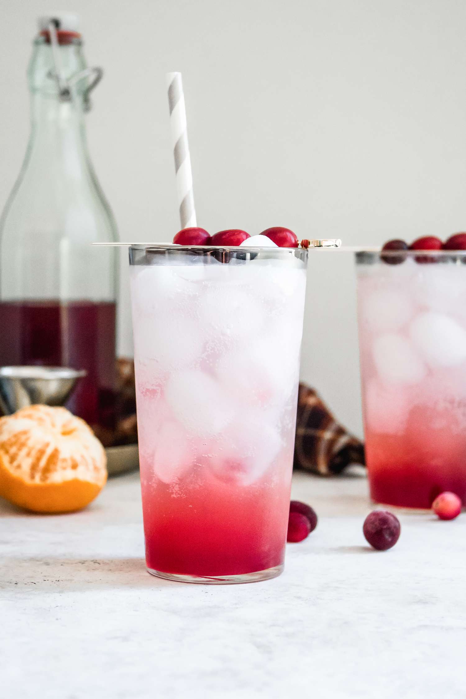 Cranberry Tangerine Shrub in a Drink Topped with Ice, Sparkling Water, and Cranberries, and in the Background, a Bottle of Shrub, Cocktail Supplies, a Peeled Tangerine, and Cranberries