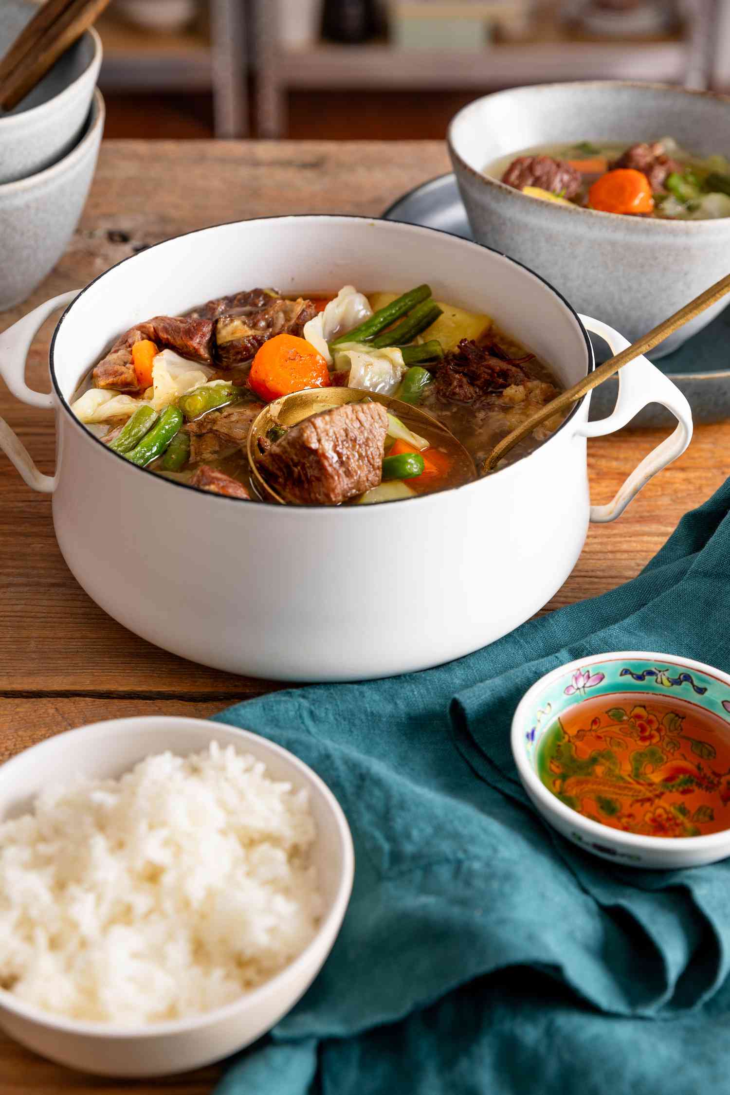 Ladle raising some nilagang baka from a Dutch oven with more, and in the surroundings, a stack of bowls, a serving of Filipino beef soup in a bowl, a bowl of rice, a small bowl of fish sauce, and a navy table napkin