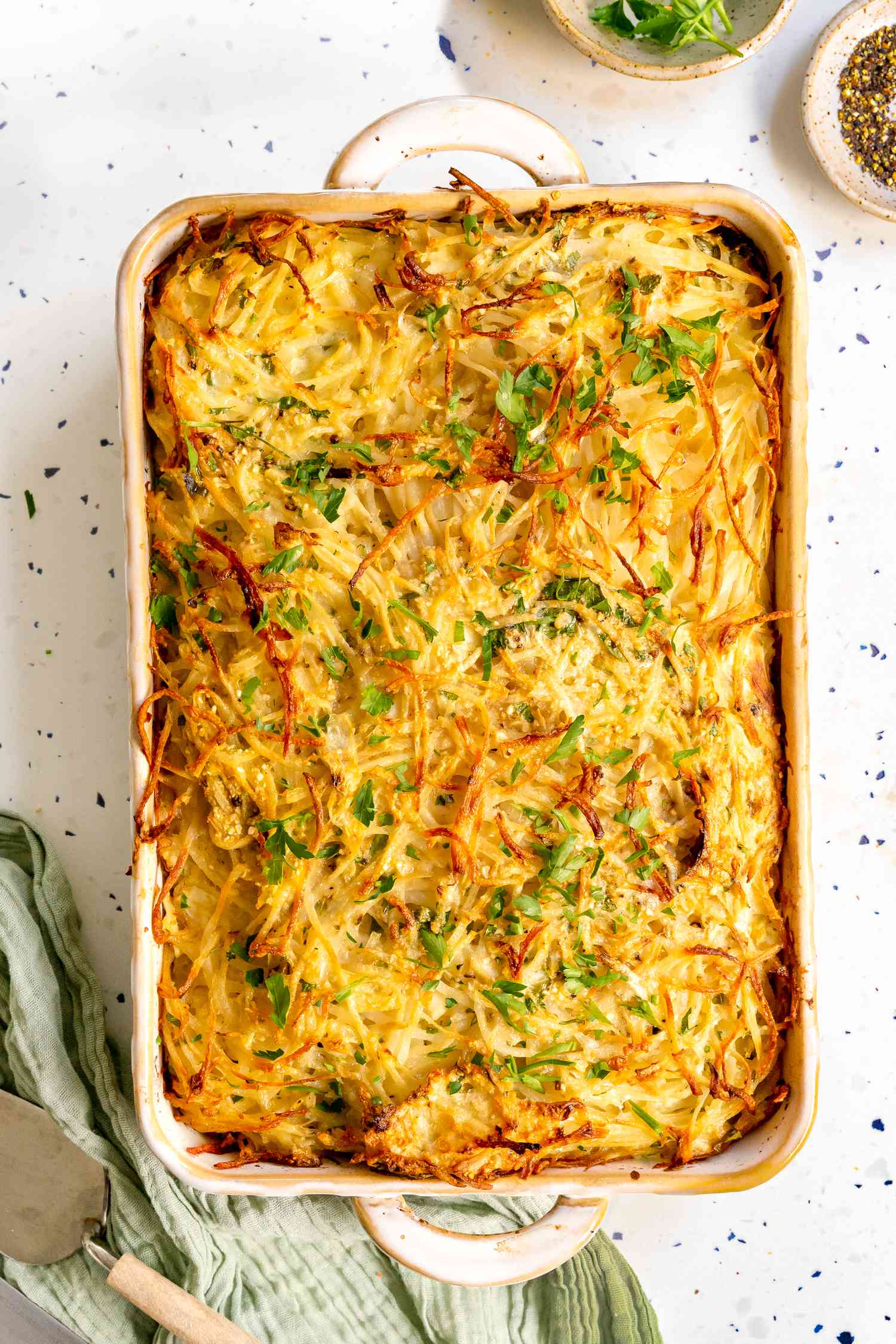 Potato Kugel in a Casserole Dish Surrounded by a Small Saucer with Herbs, Another Small Saucer with Cracked Pepper, and a Kitchen Linen
