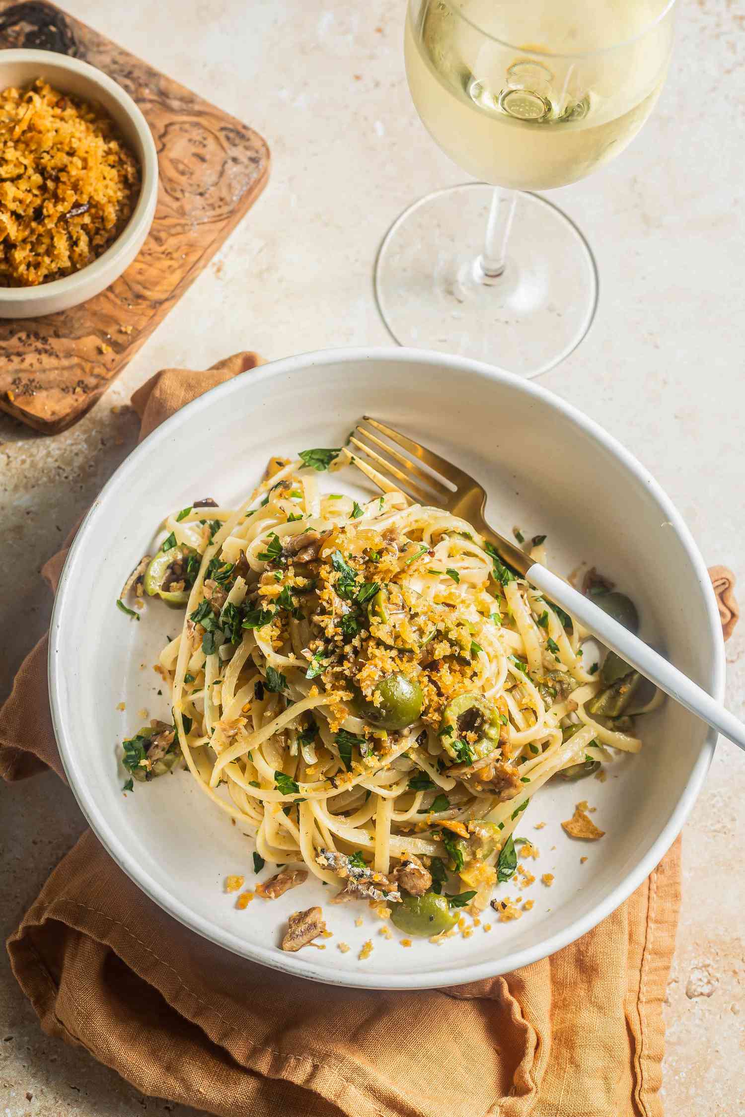 Lemony Sardine Pasta Topped With Toasted Breadcrumbs in a Bowl Next to a Glass of Wine and a Bowl With More Breadcrumbs on a Piece of Wood