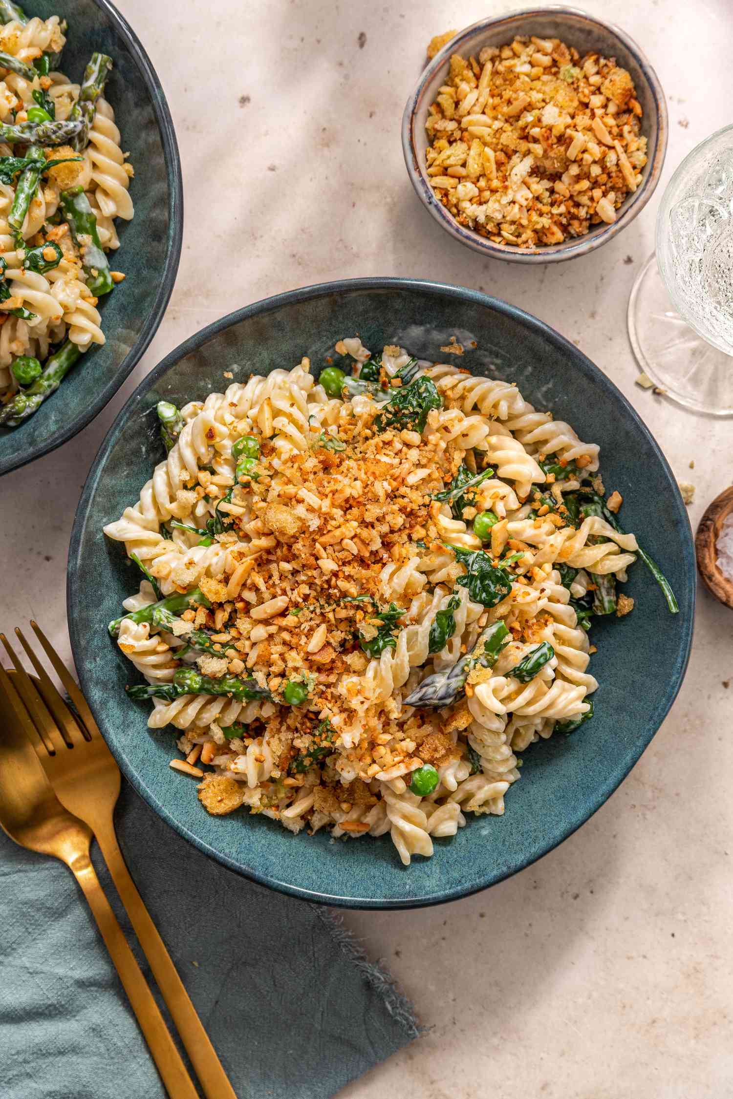 Bowl of Fusilli With Spring Vegetables and Breadcrumbs Next to Another Bowl of Pasta, a Bowl of Bread Crumb Topping, a Glass, and a Pair of Utensils on a Table Linen