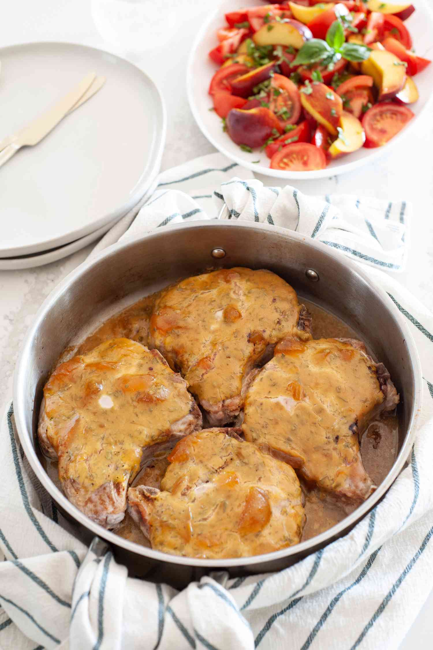 Peach Dijon Pork Chops in a Pan Next to Bowl of Tomatoes and Peaches and Plats with Utensils