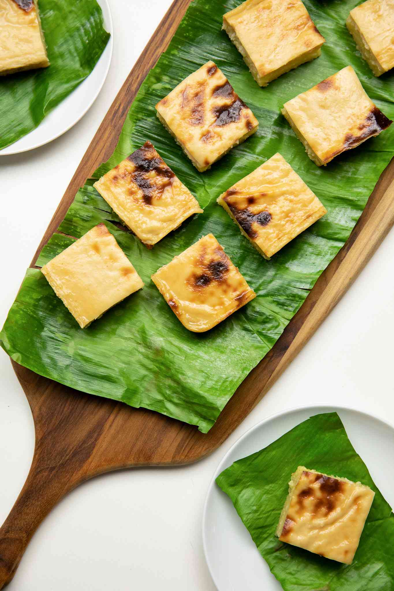 Aerial Shot: Cassava Cake Slices on a Banana Leaf Covered Wooden Tray, and Individual Slices of Cake on Banana Leaf Covered Plates