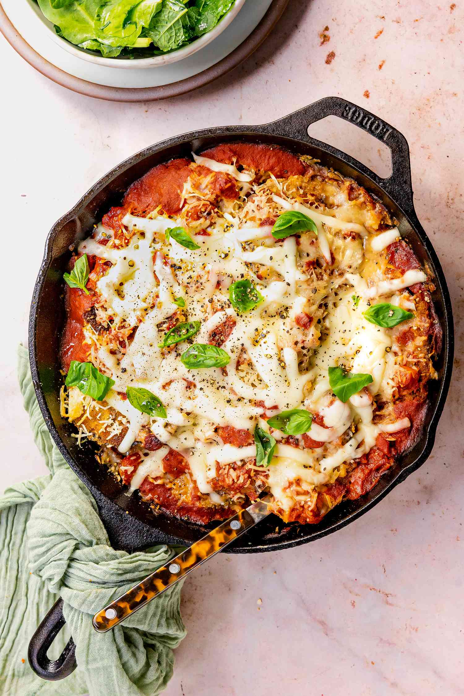 Skillet Eggplant Parmesan Topped With Fresh Basil in a Cast Iron Skillet With a Spoon, and in the Surroundings, a Bowl of Salad Greens and a Mint Kitchen Linen on the Handle of the Cast Iron Skillet