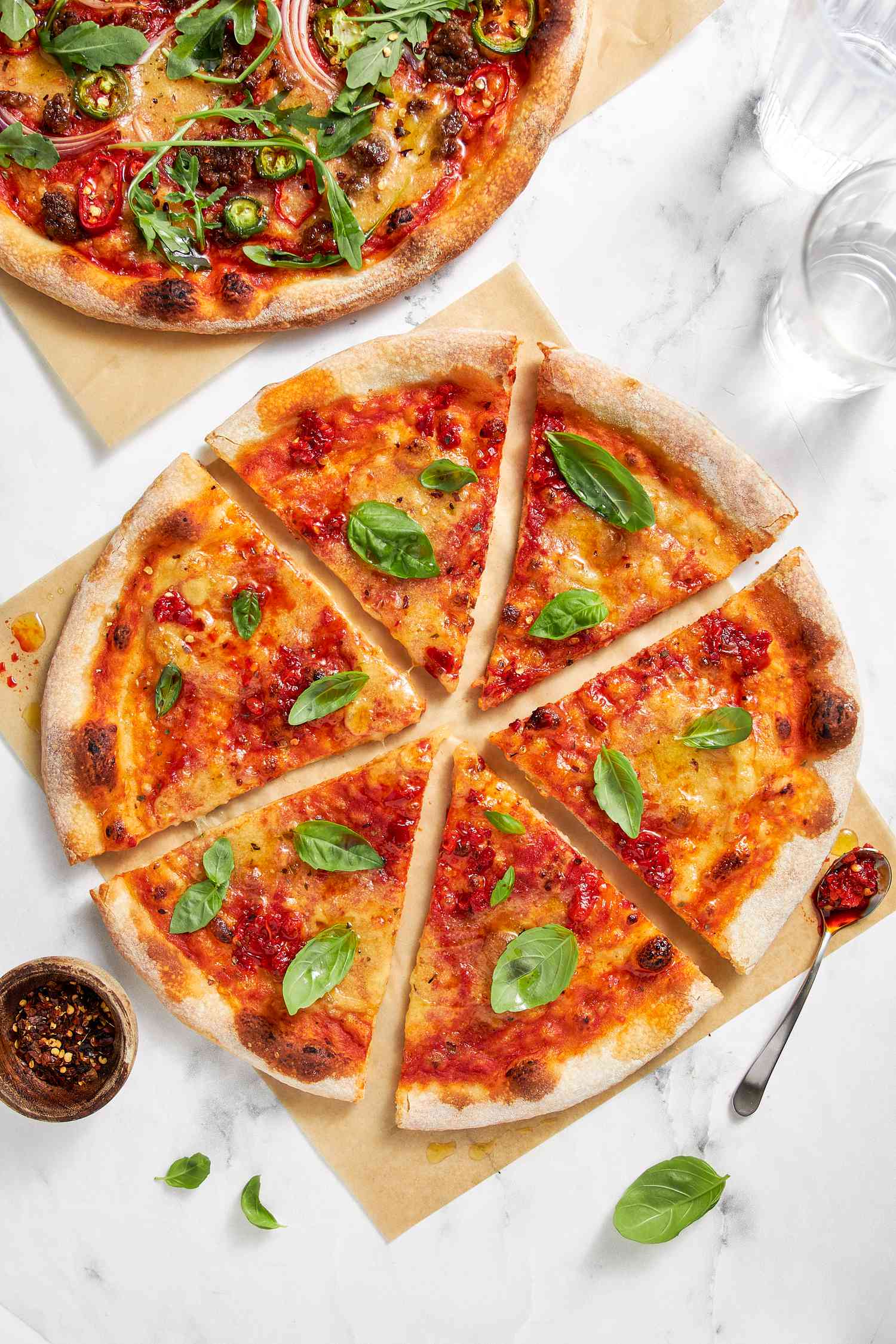 Vegan Pizza Topped With Fresh Basil and Cut Into Slices (on One Piece of Parchment Paper), Next to It, Another Vegan Pizza Topped With Jalapeno and Red Onion Slices, Arugula, and Chili Flakes (on Another Piece of Parchment Paper), a Bowl of Chili Flakes, and a Few Glasses of Water on the Counter