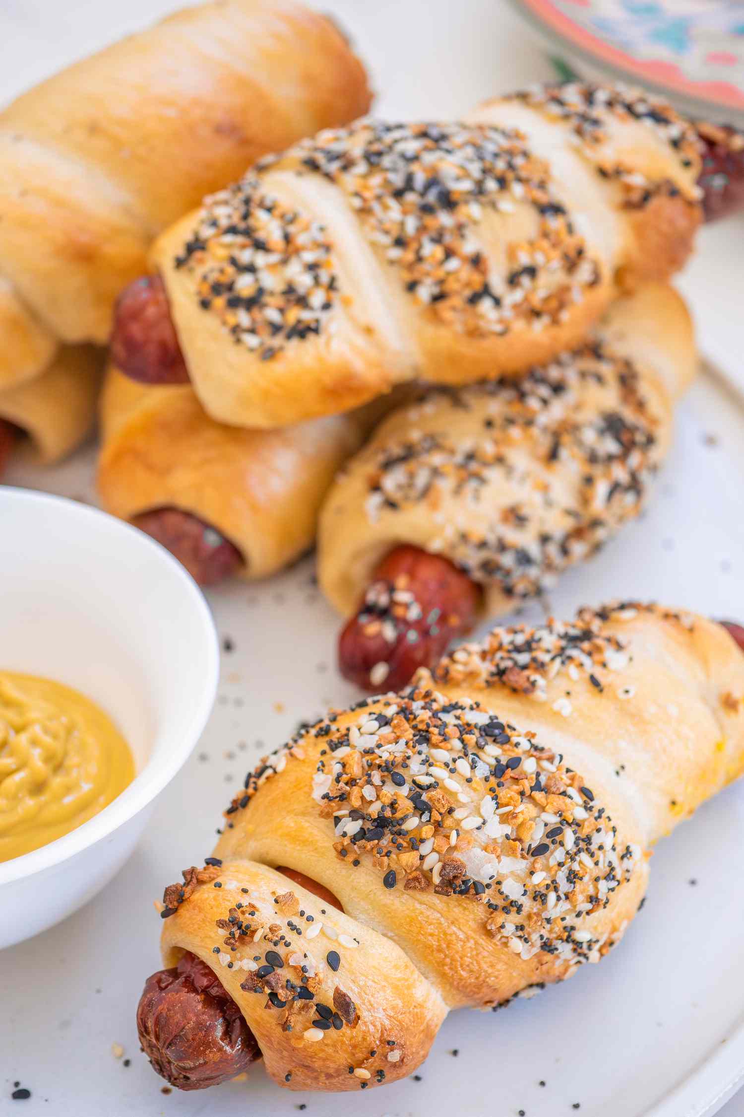 Homemade Hot Dog Bagels (Some With Everything Bagel Topping and Some Plain) on a Plate, and in the Surroundings, a Stack of Plates and a Bowl of Yellow Mustard 