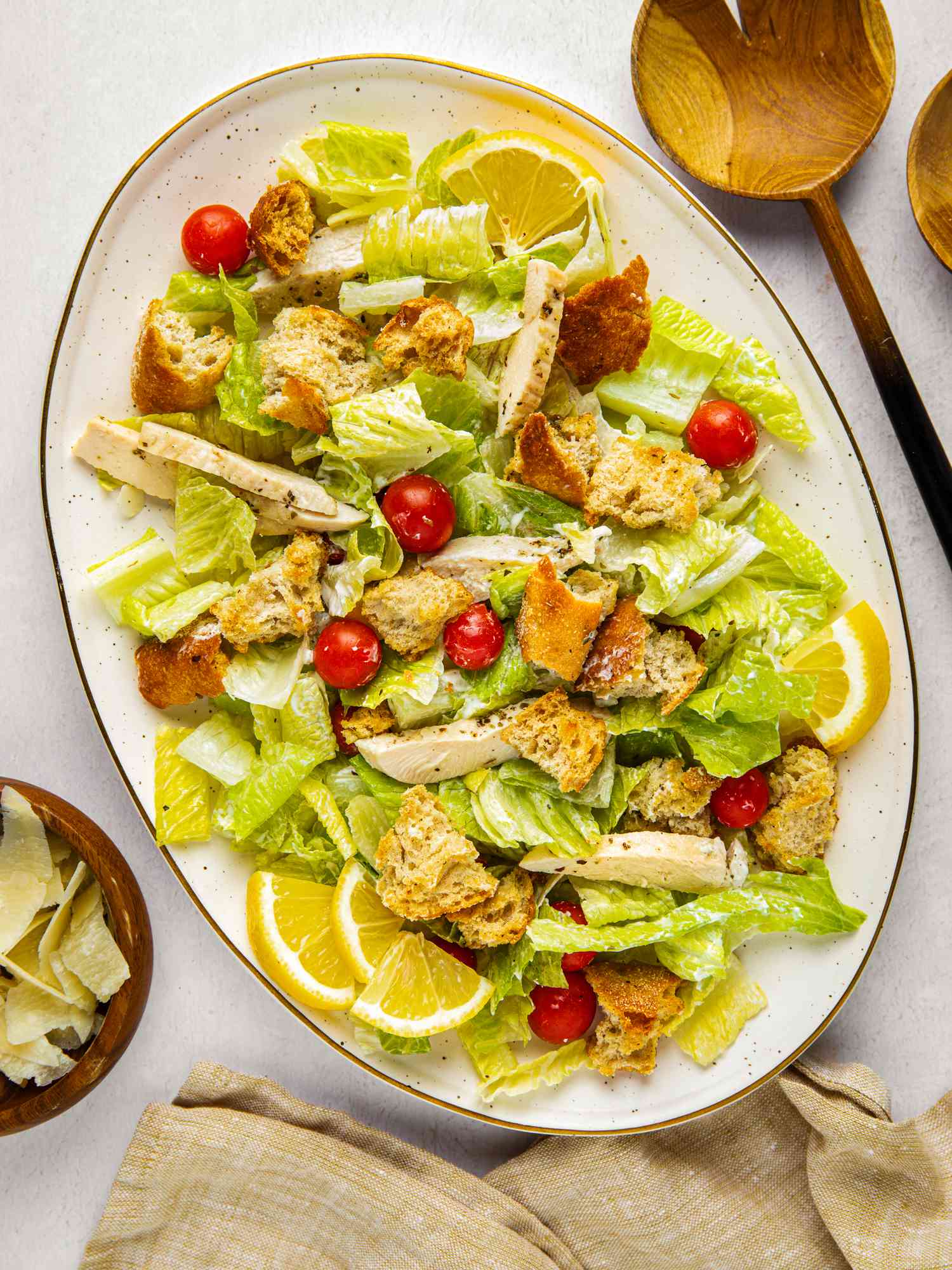 Sheet pan caesar salad on a platter with a bowl of fresh parmesan, salad utensils, and a beige table napkin