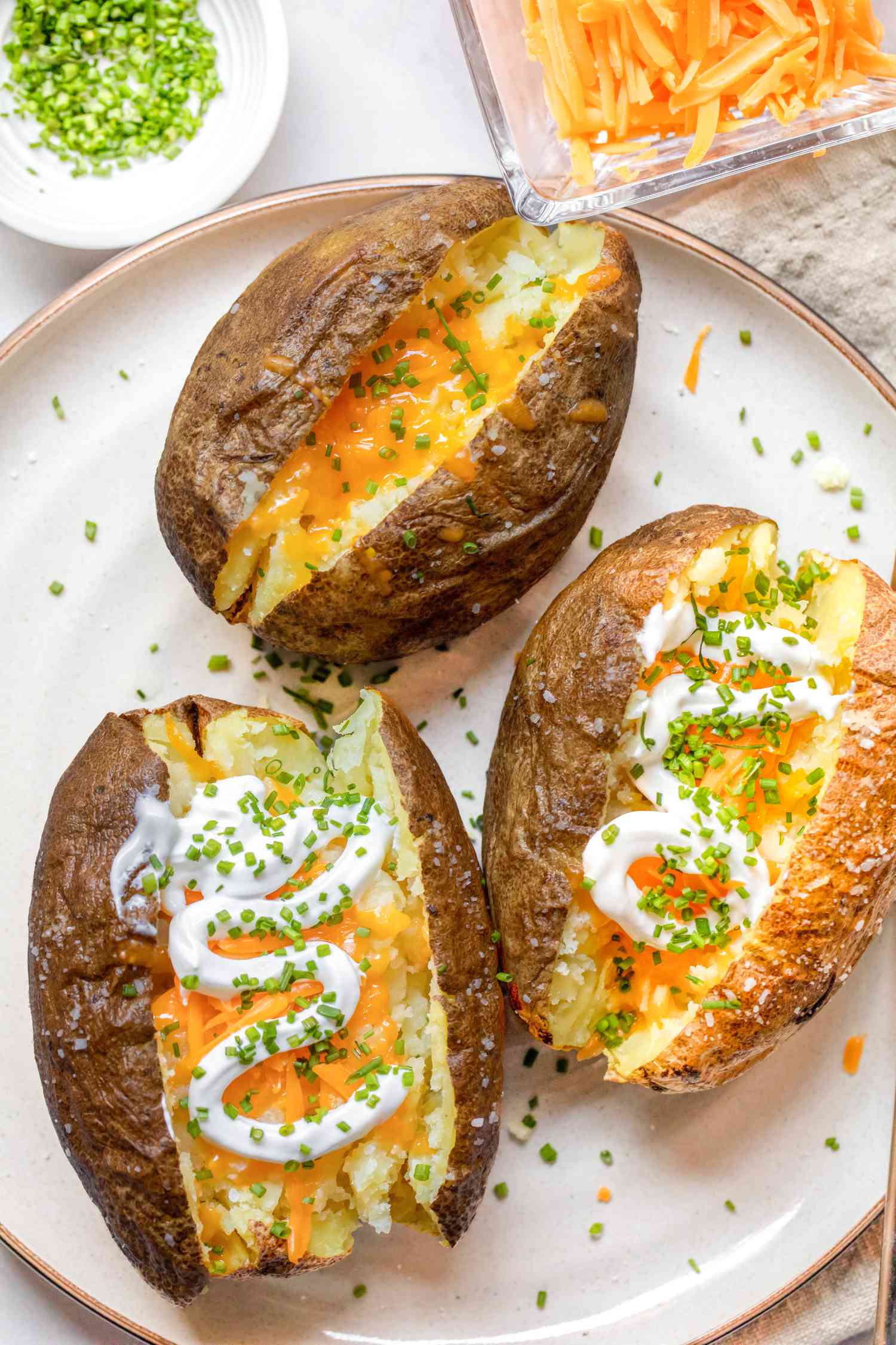 Microwave Baked Potatoes Topped with Sour Cream, Cheddar Cheese, and Chives on a Plate, and in the Background, a Small Plate with More Cheese and a Saucer with More Chives