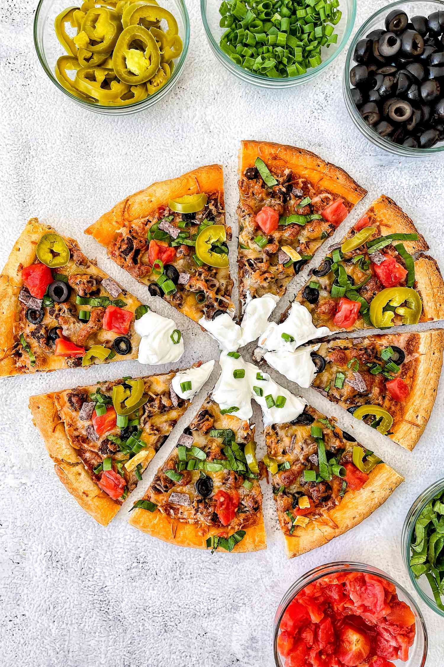 Taco Pizza on the Counter Sliced Into Pieces, and in the Surroundings, Bowls of Toppings (Sliced Scallions, Salsa, Sliced Lettuce, Sliced Olives, and Sliced JalapeÃ±os)