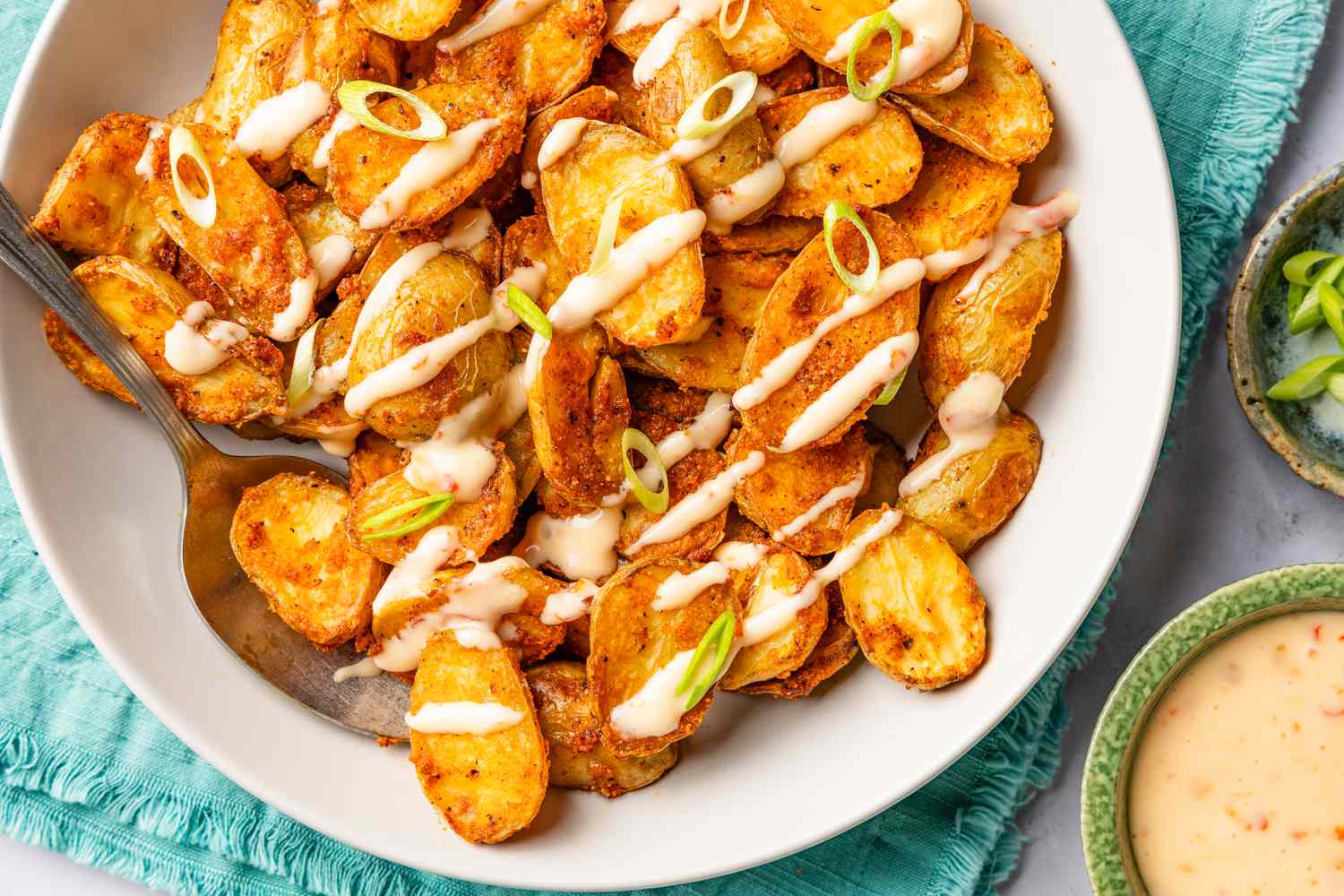 bang bang potatoes topped with sauce in a bowl at a table setting next to small bowl with more sauce and a bowl with sliced scallions