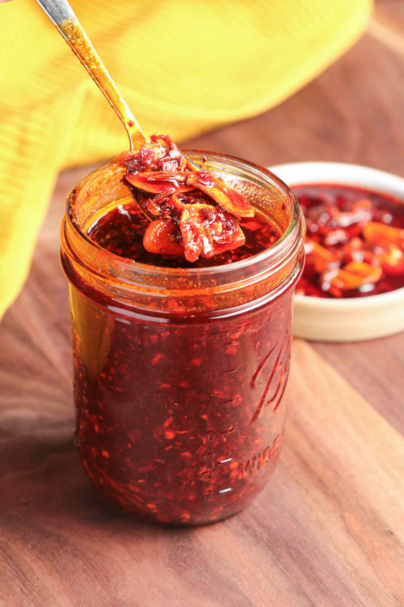 Mason Jar Filled With Chili Crisp, and in the Background, a Saucer With Some Homemade Chili Crisp and a Mustard Kitchen Towel